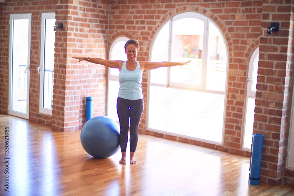Middle age beautiful sportswoman smiling happy. Streching using fitness ball in a class of yoga at gym Middle age beautiful sportswoman smiling happy. Streching using fitness ball in a class of yoga at gym