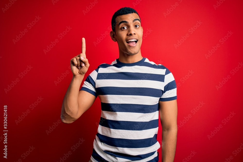 Fototapeta premium Handsome african american man wearing casual striped t-shirt standing over red background pointing finger up with successful idea. Exited and happy. Number one.