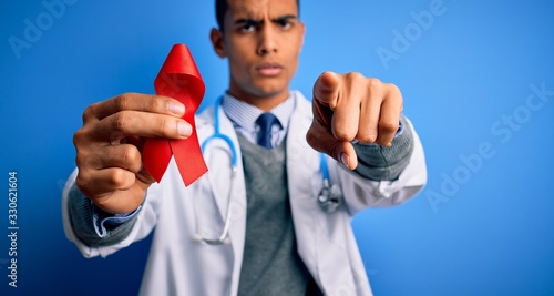 Young handsome african american doctor man holding red HIV ribbon symbol pointing with finger to the camera and to you, hand sign, positive and confident gesture from the front