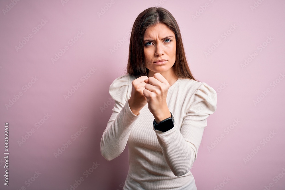 Young beautiful woman with blue eyes wearing casual white t-shirt over pink background Ready to fight with fist defense gesture, angry and upset face, afraid of problem Young beautiful woman with blue eyes wearing casual white t-shirt over pink background Ready to fight with fist defense gesture, angry and upset face, afraid of problem
