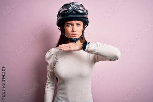 Young beautiful motorcyclist woman with blue eyes wearing moto helmet over pink background cutting throat with hand as knife, threaten aggression with furious violence