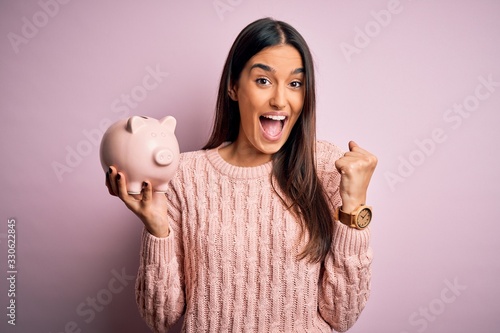 Young beautiful brunette woman holding piggy bank saving money for retirement screaming proud and celebrating victory and success very excited, cheering emotion photo