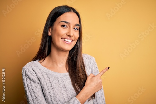 Young beautiful brunette woman wearing white casual sweater over yellow background cheerful with a smile of face pointing with hand and finger up to the side with happy and natural expression on face