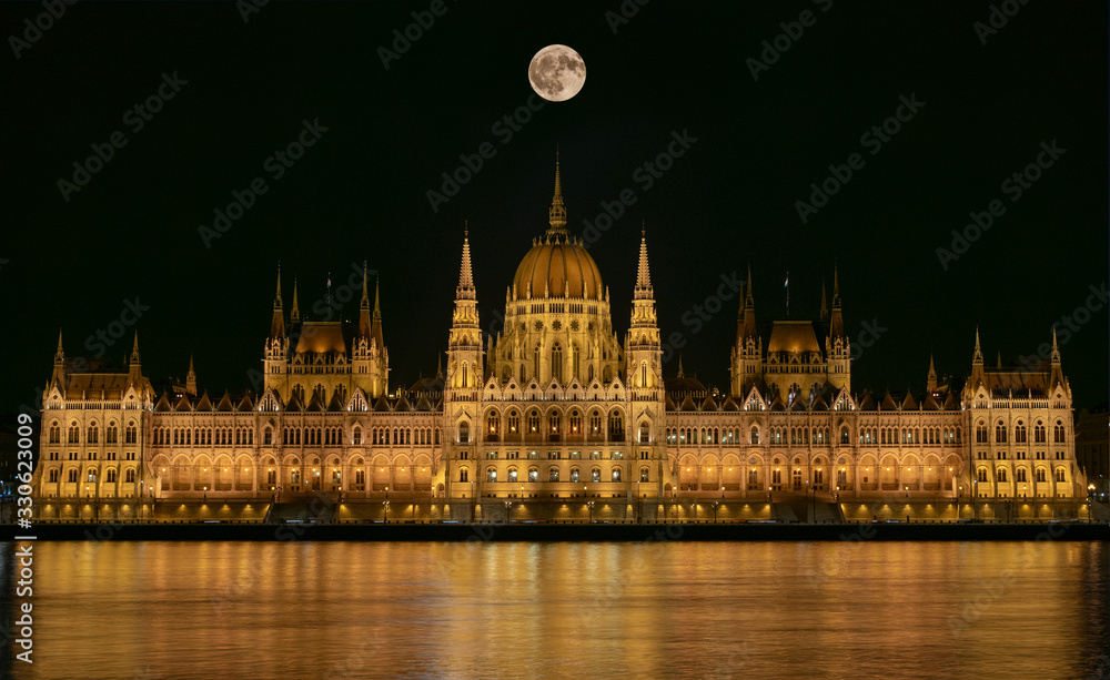 Fototapeta premium Budapest Parliament Building illuminated at night with dark sky, giant full moon, reflection in Danube river and river with silk texture.