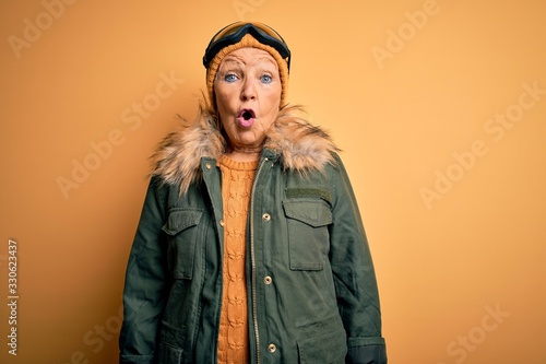 Senior beautiful grey-haired skier woman wearing snow sportswear and ski goggles afraid and shocked with surprise expression, fear and excited face.