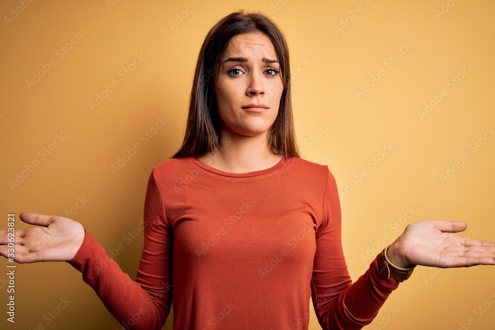 Obraz premium Young beautiful brunette woman wearing casual t-shirt standing over yellow background clueless and confused with open arms, no idea concept.