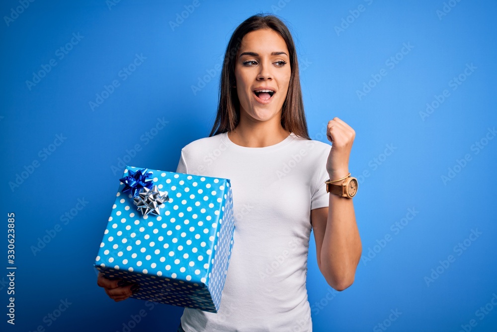 Young beautiful brunette woman holding birthday gift over isolated blue background pointing and showing with thumb up to the side with happy face smiling Young beautiful brunette woman holding birthday gift over isolated blue background pointing and showing with thumb up to the side with happy face smiling
