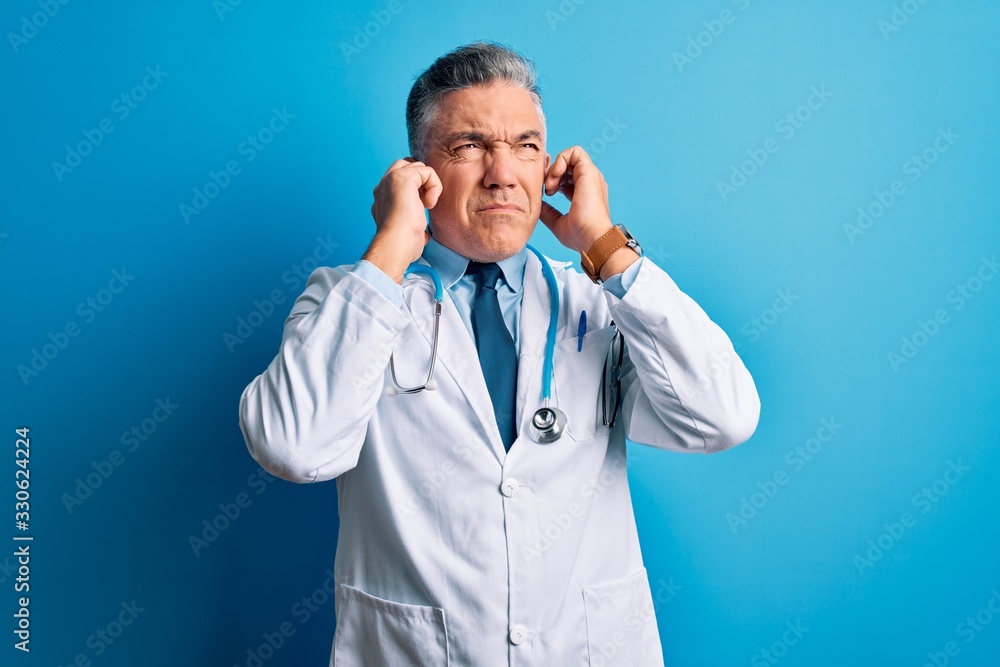 Middle age handsome grey-haired doctor man wearing coat and blue stethoscope covering ears with fingers with annoyed expression for the noise of loud music. Deaf concept. Middle age handsome grey-haired doctor man wearing coat and blue stethoscope covering ears with fingers with annoyed expression for the noise of loud music. Deaf concept.