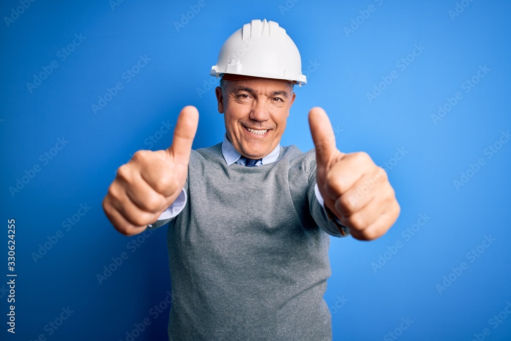 Middle age handsome grey-haired engineer man wearing safety helmet over blue background approving doing positive gesture with hand, thumbs up smiling and happy for success. Winner gesture. Middle age handsome grey-haired engineer man wearing safety helmet over blue background approving doing positive gesture with hand, thumbs up smiling and happy for success. Winner gesture.