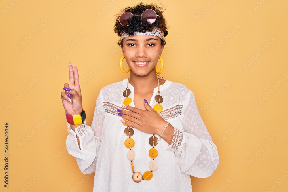 Young beautiful african american afro hippie woman wearing sunglasses and accessories smiling swearing with hand on chest and fingers up, making a loyalty promise oath