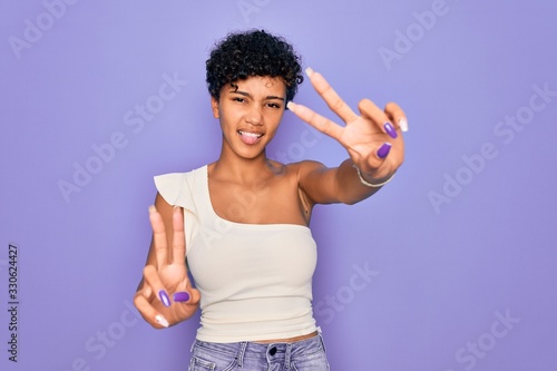 Young beautiful african american afro woman wearing casual t-shirt over purple background smiling with tongue out showing fingers of both hands doing victory sign. Number two.