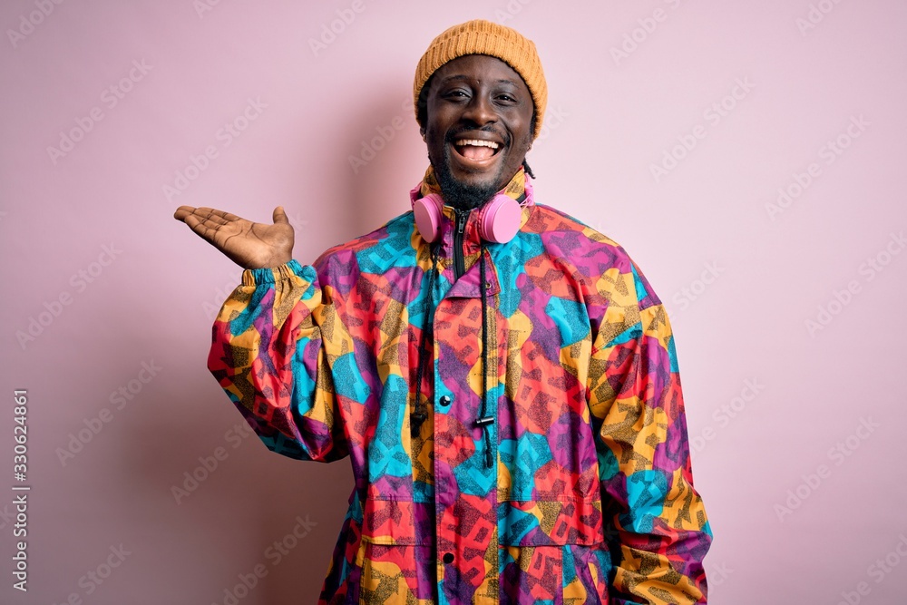 Young handsome african american man wearing colorful coat and cap over pink background smiling cheerful presenting and pointing with palm of hand looking at the camera.