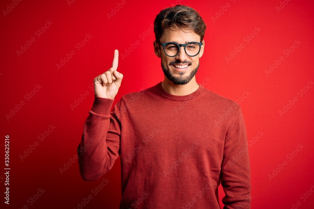 Young handsome man with beard wearing glasses and sweater standing over red background showing and pointing up with finger number one while smiling confident and happy. Young handsome man with beard wearing glasses and sweater standing over red background showing and pointing up with finger number one while smiling confident and happy.