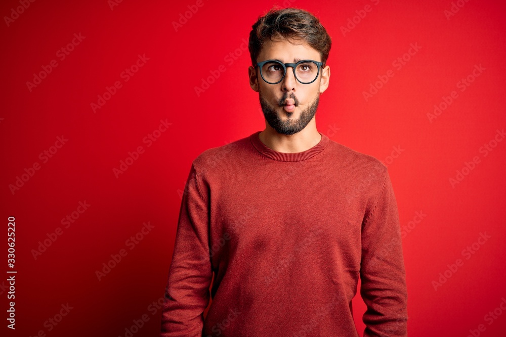 Young handsome man with beard wearing glasses and sweater standing over red background making fish face with lips, crazy and comical gesture. Funny expression. Young handsome man with beard wearing glasses and sweater standing over red background making fish face with lips, crazy and comical gesture. Funny expression.