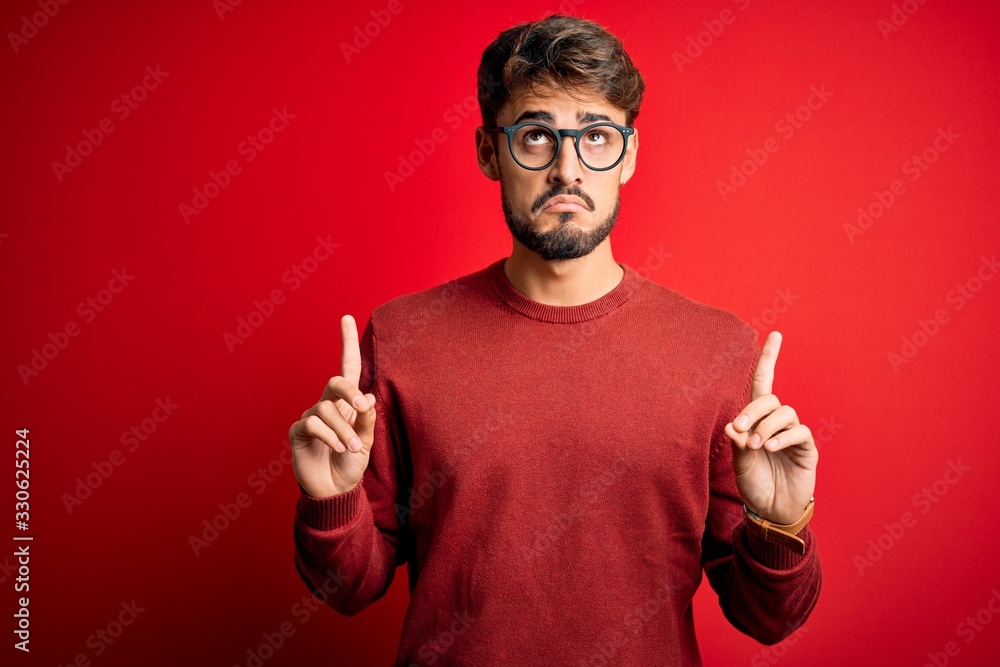 Young handsome man with beard wearing glasses and sweater standing over red background Pointing up looking sad and upset, indicating direction with fingers, unhappy and depressed. Young handsome man with beard wearing glasses and sweater standing over red background Pointing up looking sad and upset, indicating direction with fingers, unhappy and depressed.