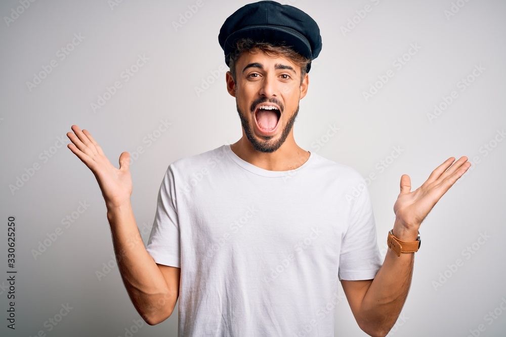 Young driver man with beard wearing hat standing over isolated white background celebrating crazy and amazed for success with arms raised and open eyes screaming excited. Winner concept Young driver man with beard wearing hat standing over isolated white background celebrating crazy and amazed for success with arms raised and open eyes screaming excited. Winner concept