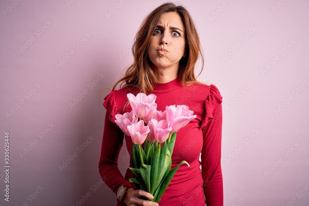 Young beautiful brunette woman holding bouquet of pink tulips over isolated background puffing cheeks with funny face. Mouth inflated with air, crazy expression. Young beautiful brunette woman holding bouquet of pink tulips over isolated background puffing cheeks with funny face. Mouth inflated with air, crazy expression.