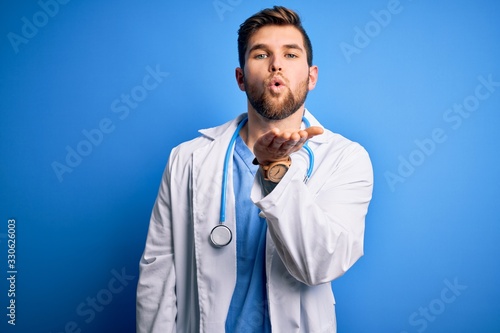 Young blond doctor man with beard and blue eyes wearing white coat and stethoscope looking at the camera blowing a kiss with hand on air being lovely and sexy. Love expression.