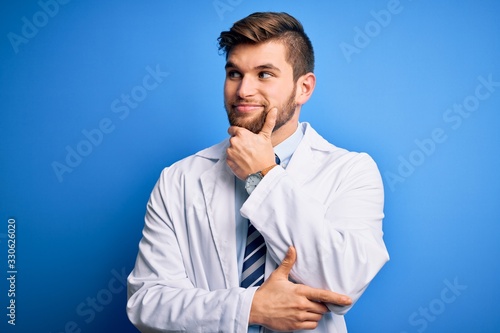 Young blond therapist man with beard and blue eyes wearing coat and tie over background with hand on chin thinking about question, pensive expression. Smiling with thoughtful face. Doubt concept.