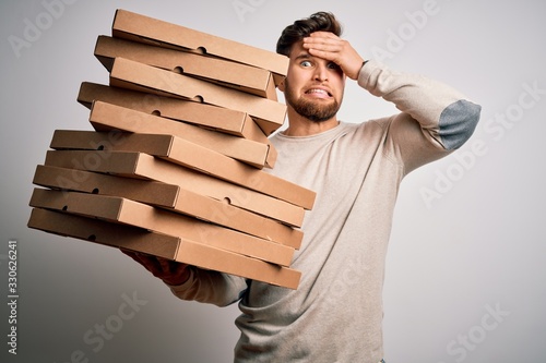 Young blond man with beard and blue eyes holding cardboards of pizza over white background stressed with hand on head, shocked with shame and surprise face, angry and frustrated. Fear and upset