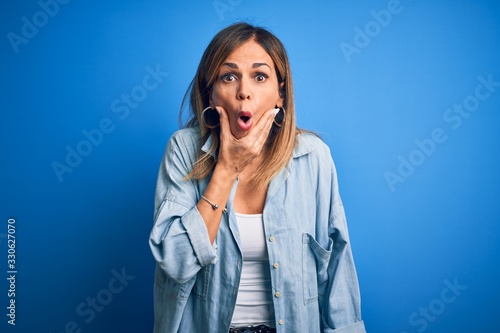 Middle age beautiful woman wearing casual shirt standing over isolated blue background Looking fascinated with disbelief, surprise and amazed expression with hands on chin