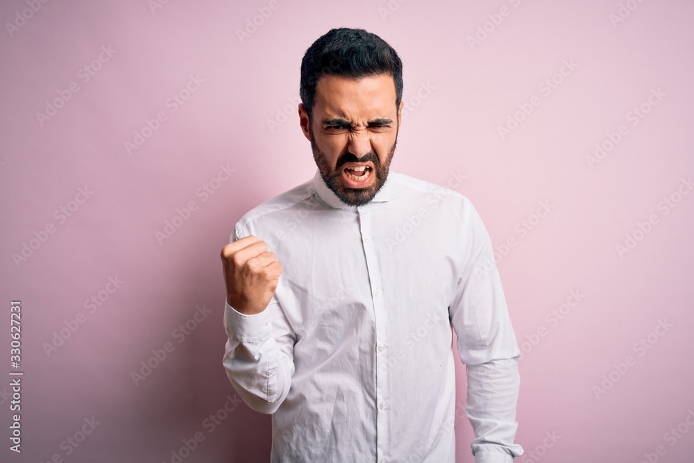 Young handsome man with beard wearing casual shirt standing over pink background angry and mad raising fist frustrated and furious while shouting with anger. Rage and aggressive concept. Young handsome man with beard wearing casual shirt standing over pink background angry and mad raising fist frustrated and furious while shouting with anger. Rage and aggressive concept.