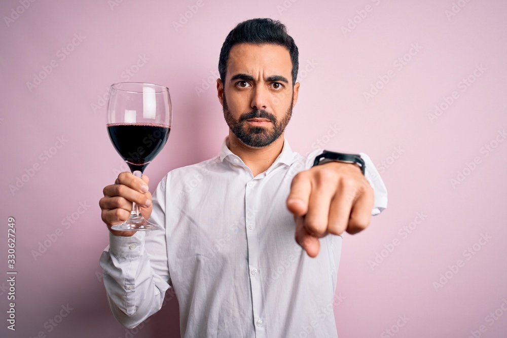 Young handsome man with beard drinking glass of red wine over isolated pink background pointing with finger to the camera and to you, hand sign, positive and confident gesture from the front Young handsome man with beard drinking glass of red wine over isolated pink background pointing with finger to the camera and to you, hand sign, positive and confident gesture from the front