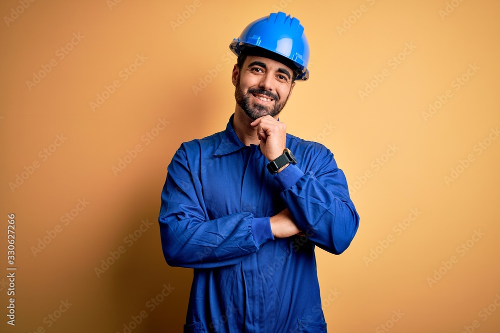 Mechanic man with beard wearing blue uniform and safety helmet over yellow background looking confident at the camera smiling with crossed arms and hand raised on chin. Thinking positive.