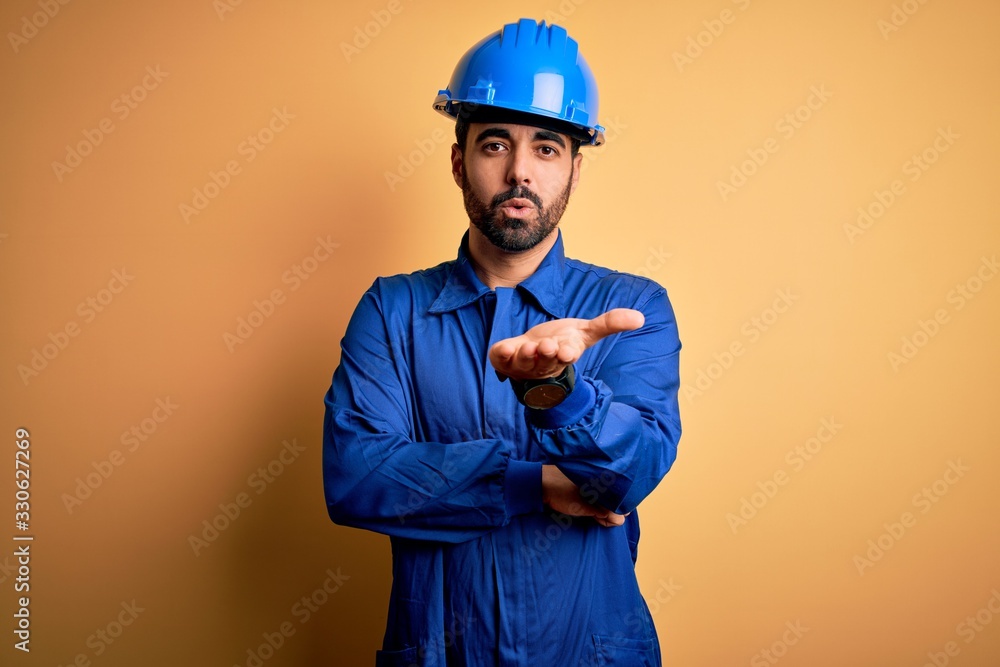 Mechanic man with beard wearing blue uniform and safety helmet over yellow background looking at the camera blowing a kiss with hand on air being lovely and sexy. Love expression.