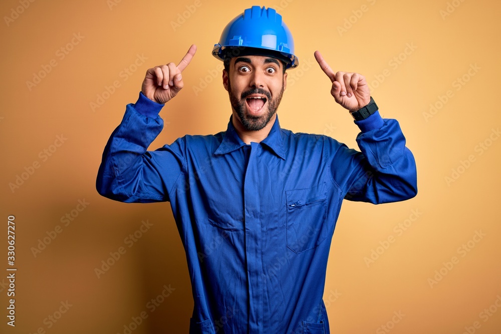 Mechanic man with beard wearing blue uniform and safety helmet over yellow background smiling amazed and surprised and pointing up with fingers and raised arms.