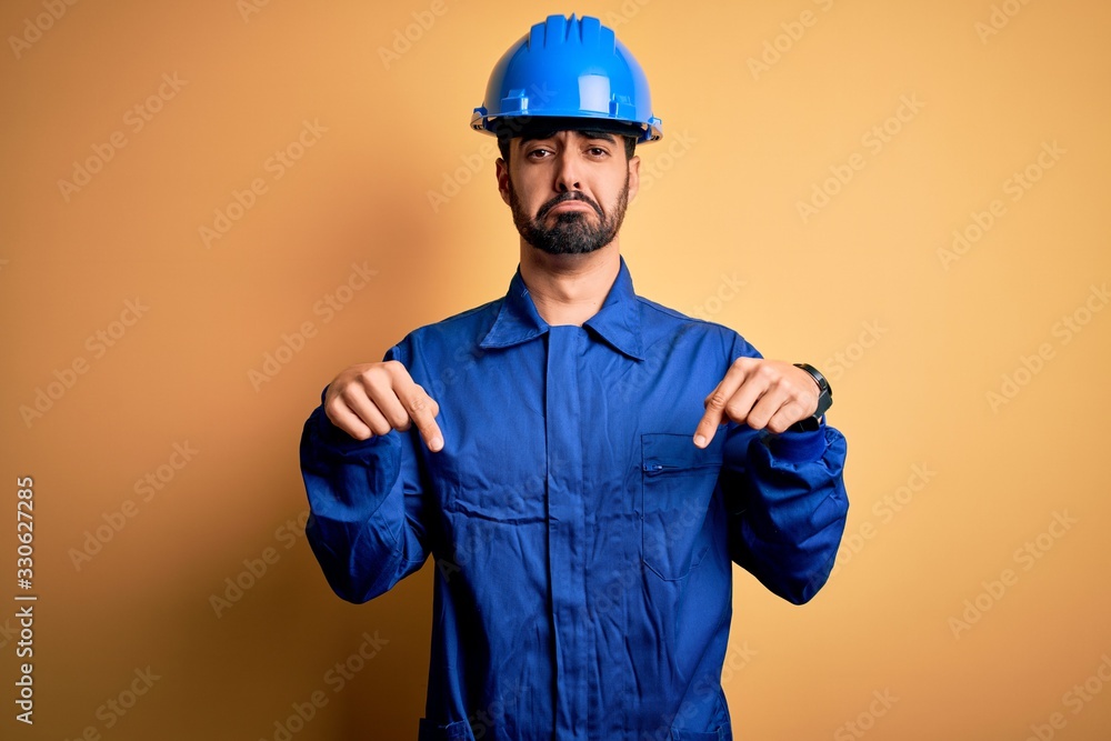Mechanic man with beard wearing blue uniform and safety helmet over yellow background Pointing down looking sad and upset, indicating direction with fingers, unhappy and depressed.