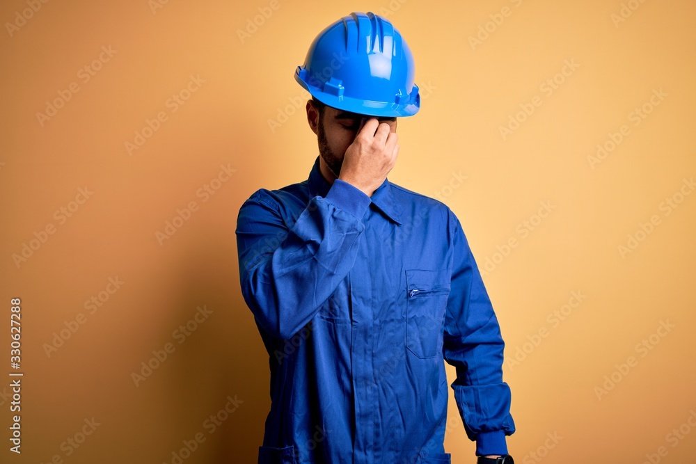 Mechanic man with beard wearing blue uniform and safety helmet over yellow background tired rubbing nose and eyes feeling fatigue and headache. Stress and frustration concept.