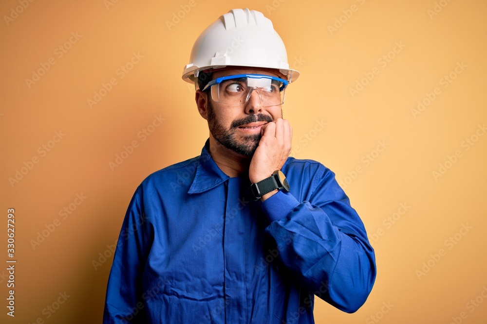 Mechanic man with beard wearing blue uniform and safety glasses over yellow background looking stressed and nervous with hands on mouth biting nails. Anxiety problem.