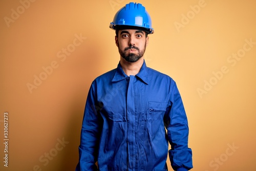 Mechanic man with beard wearing blue uniform and safety helmet over yellow background puffing cheeks with funny face. Mouth inflated with air, crazy expression.