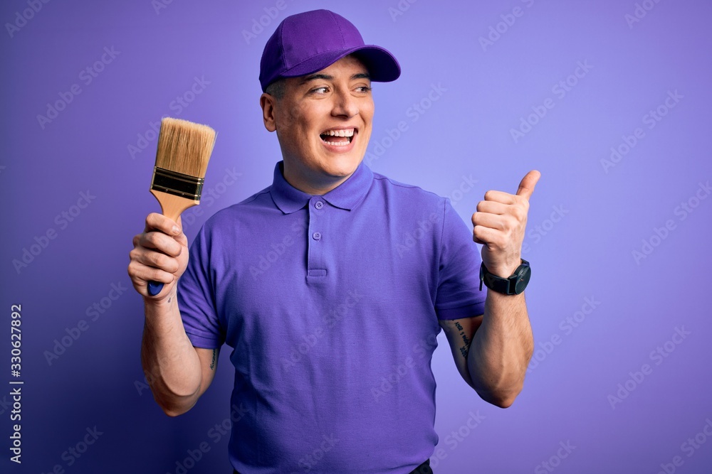Young modern decorator painter man holding paint brush over purple background pointing and showing with thumb up to the side with happy face smiling Young modern decorator painter man holding paint brush over purple background pointing and showing with thumb up to the side with happy face smiling