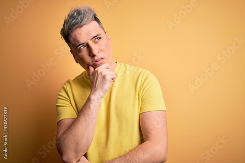 Young handsome modern man wearing yellow shirt over yellow isolated background with hand on chin thinking about question, pensive expression. Smiling with thoughtful face. Doubt concept.