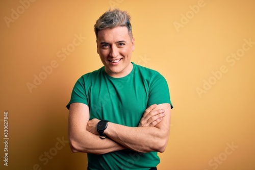 Young handsome modern man wearing casual green t-shirt over yellow background happy face smiling with crossed arms looking at the camera. Positive person.