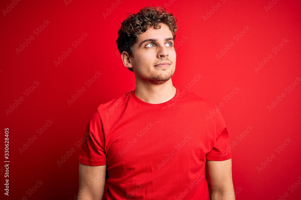Young blond handsome man with curly hair wearing casual t-shirt over red background smiling looking to the side and staring away thinking. Young blond handsome man with curly hair wearing casual t-shirt over red background smiling looking to the side and staring away thinking.