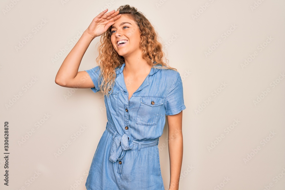 Young beautiful woman with blue eyes wearing casual denim dress over white background very happy and smiling looking far away with hand over head. Searching concept. Young beautiful woman with blue eyes wearing casual denim dress over white background very happy and smiling looking far away with hand over head. Searching concept.