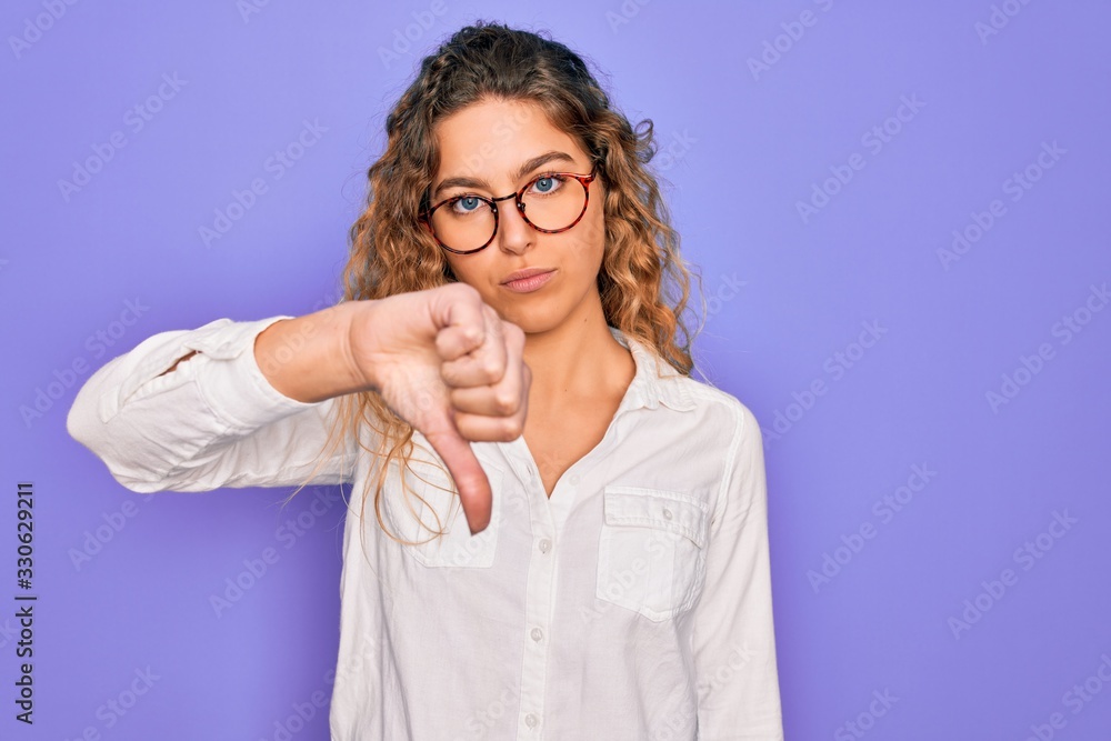 Young beautiful woman with blue eyes wearing casual shirt and glasses over purple background looking unhappy and angry showing rejection and negative with thumbs down gesture. Bad expression.