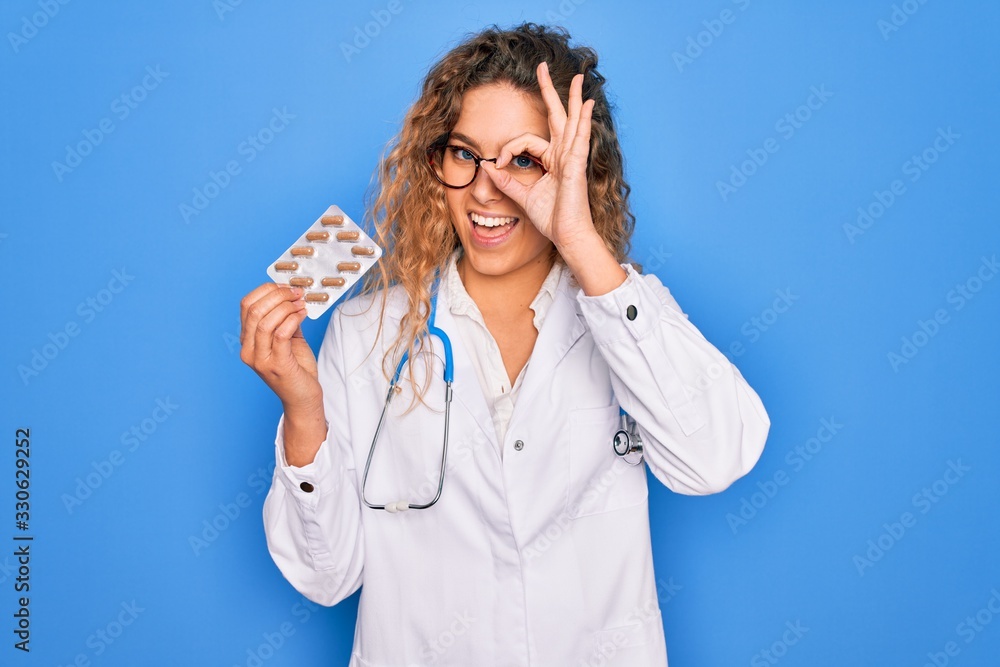 Young beautiful blonde doctor woman with blue eyes wearing stethoscope holding pills with happy face smiling doing ok sign with hand on eye looking through fingers Young beautiful blonde doctor woman with blue eyes wearing stethoscope holding pills with happy face smiling doing ok sign with hand on eye looking through fingers