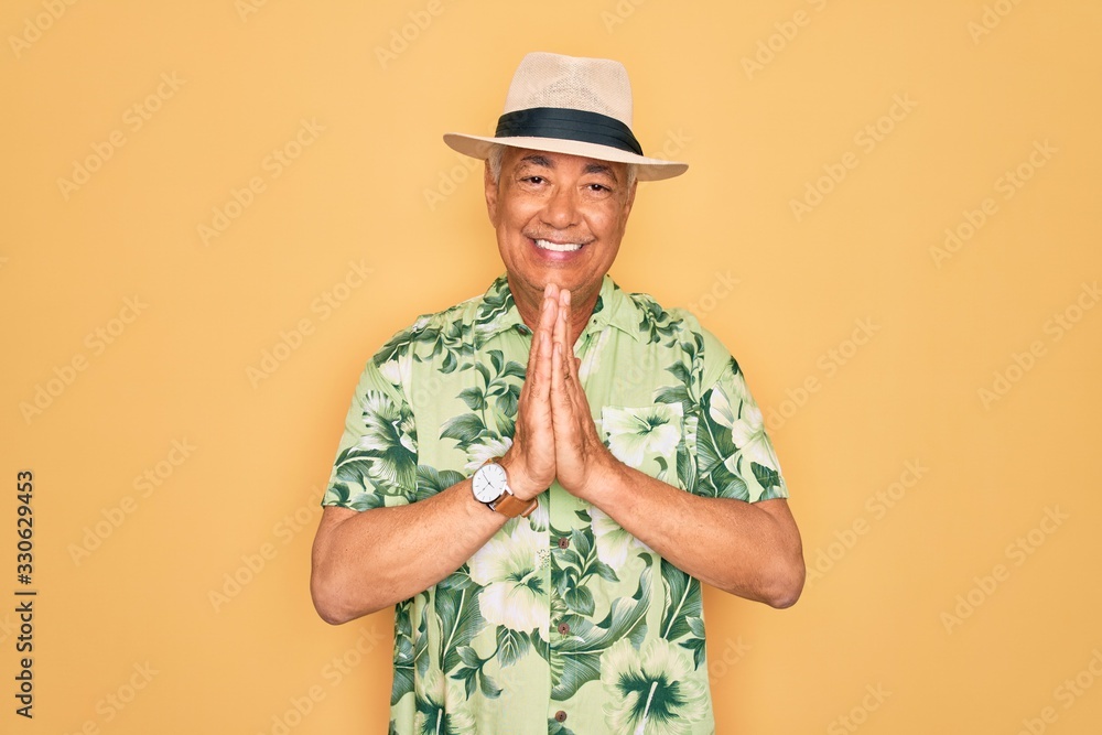 Middle age senior grey-haired man wearing summer hat and floral shirt on beach vacation praying with hands together asking for forgiveness smiling confident.