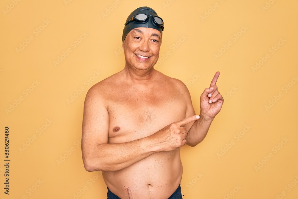 Middle age senior grey-haired swimmer man wearing swimsuit, cap and goggles smiling and looking at the camera pointing with two hands and fingers to the side.