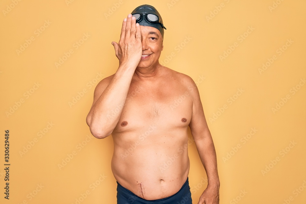 Middle age senior grey-haired swimmer man wearing swimsuit, cap and goggles covering one eye with hand, confident smile on face and surprise emotion.