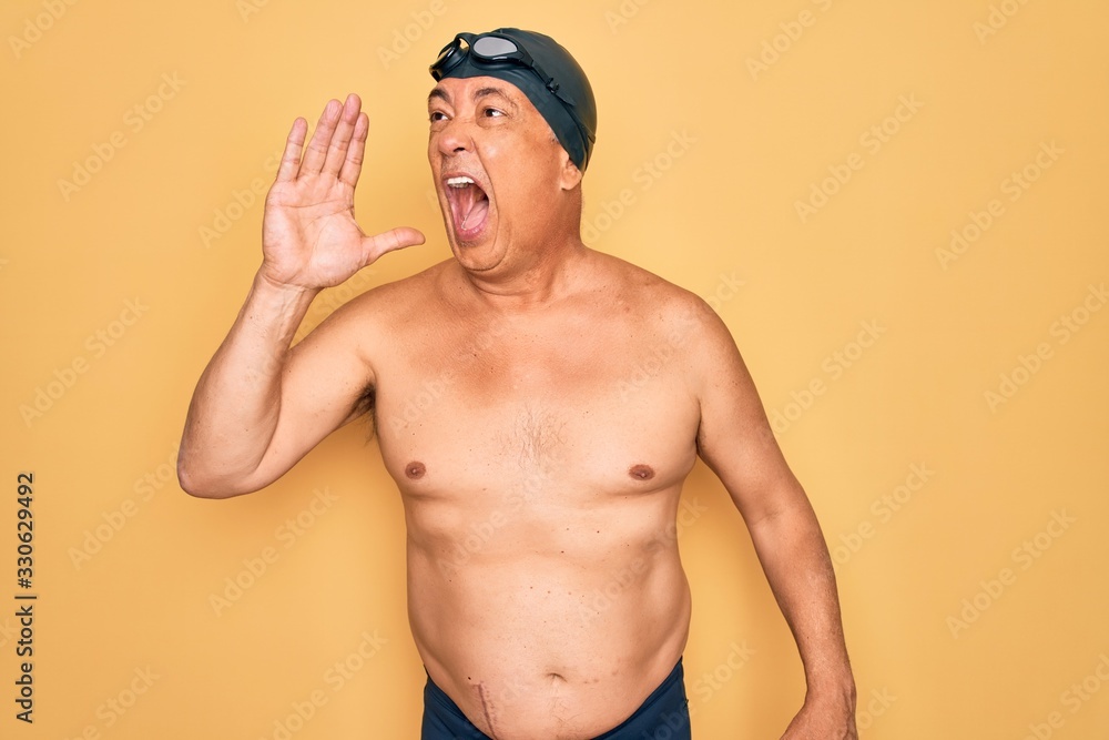 Middle age senior grey-haired swimmer man wearing swimsuit, cap and goggles shouting and screaming loud to side with hand on mouth. Communication concept.