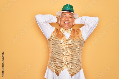 Middle age senior grey-haired man wearing Brazilian carnival custome over yellow background relaxing and stretching, arms and hands behind head and neck smiling happy photo