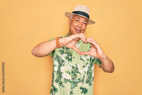 Middle age senior grey-haired man wearing summer hat and floral shirt on beach vacation smiling in love doing heart symbol shape with hands. Romantic concept.