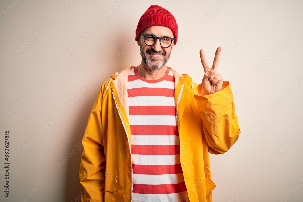 Middle age hoary man wearing glasses and rain coat standing over isolated white background smiling with happy face winking at the camera doing victory sign. Number two.