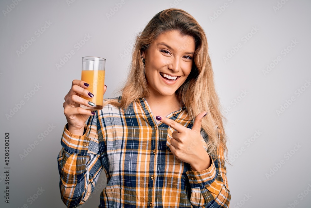 Young beautiful blonde woman drinking glass of healthy orange juice over white background very happy pointing with hand and finger Young beautiful blonde woman drinking glass of healthy orange juice over white background very happy pointing with hand and finger