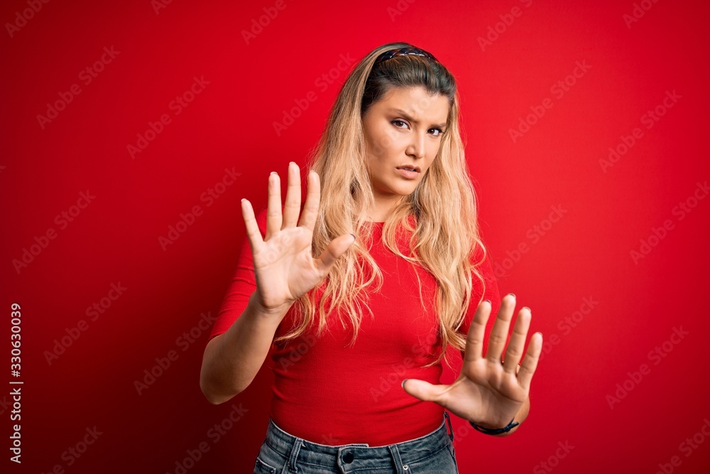 Young beautiful blonde woman wearing casual t-shirt standing over isolated red background Moving away hands palms showing refusal and denial with afraid and disgusting expression. Stop and forbidden. Young beautiful blonde woman wearing casual t-shirt standing over isolated red background Moving away hands palms showing refusal and denial with afraid and disgusting expression. Stop and forbidden.